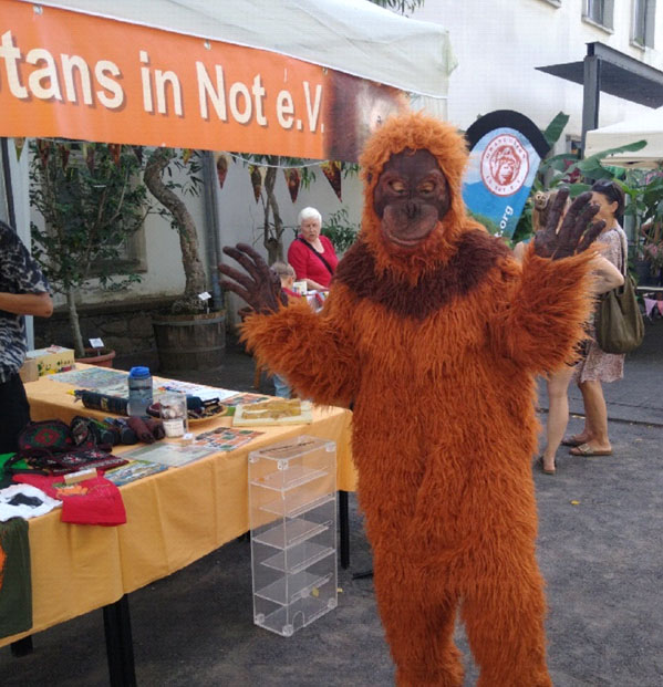 Links am Bildrand befindet sich ein Infostand vom Orang-Utans in Not e.V. In der Mitte steht ein Mensch im Orang-Utan-Kostüm, welcher die Hände in die Höhe streckt.