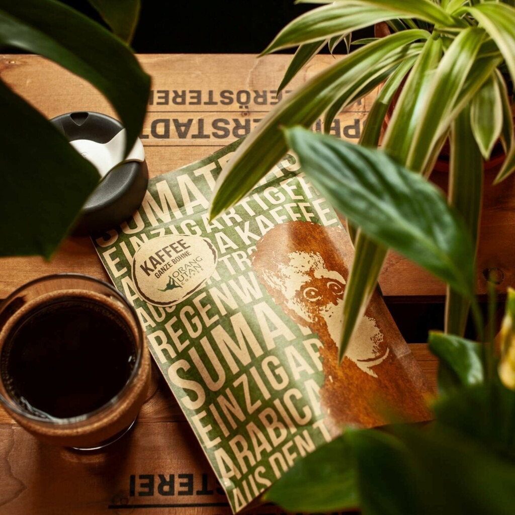 Eine Tüte des Orang-Utan-Kaffees zwischen Blättern und einem Glas mit Kaffee.