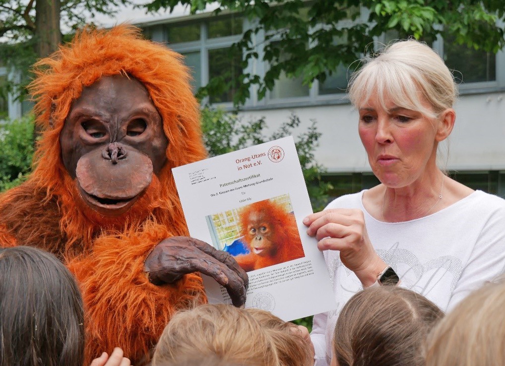 Ein Mensch in einem Orang-Utan-Kostüm überreicht ein Patenschaftszertifikat an eine Lehrerin.