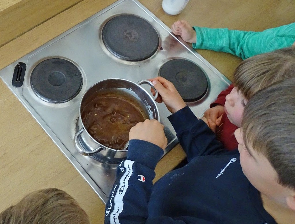 Kinder stehen am Herd und schauen in einen Topf, in dem ein Junge geschmolzene Schokolade rührt.