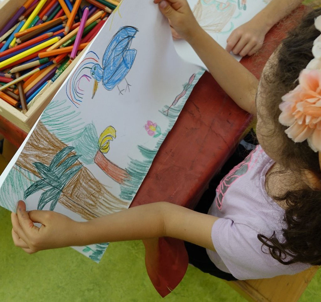 Ein Kind schaut sich ein mit Buntstiften gemaltes Bild von einem großen blauen Vogel und einigen Bäumen an.