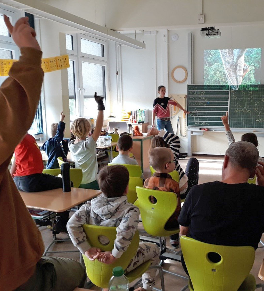 In einem Klassenzimmer befinden sich viele Kinder, einige davon melden sich. Vorn an der Tafel steht die Umweltbildnerin.