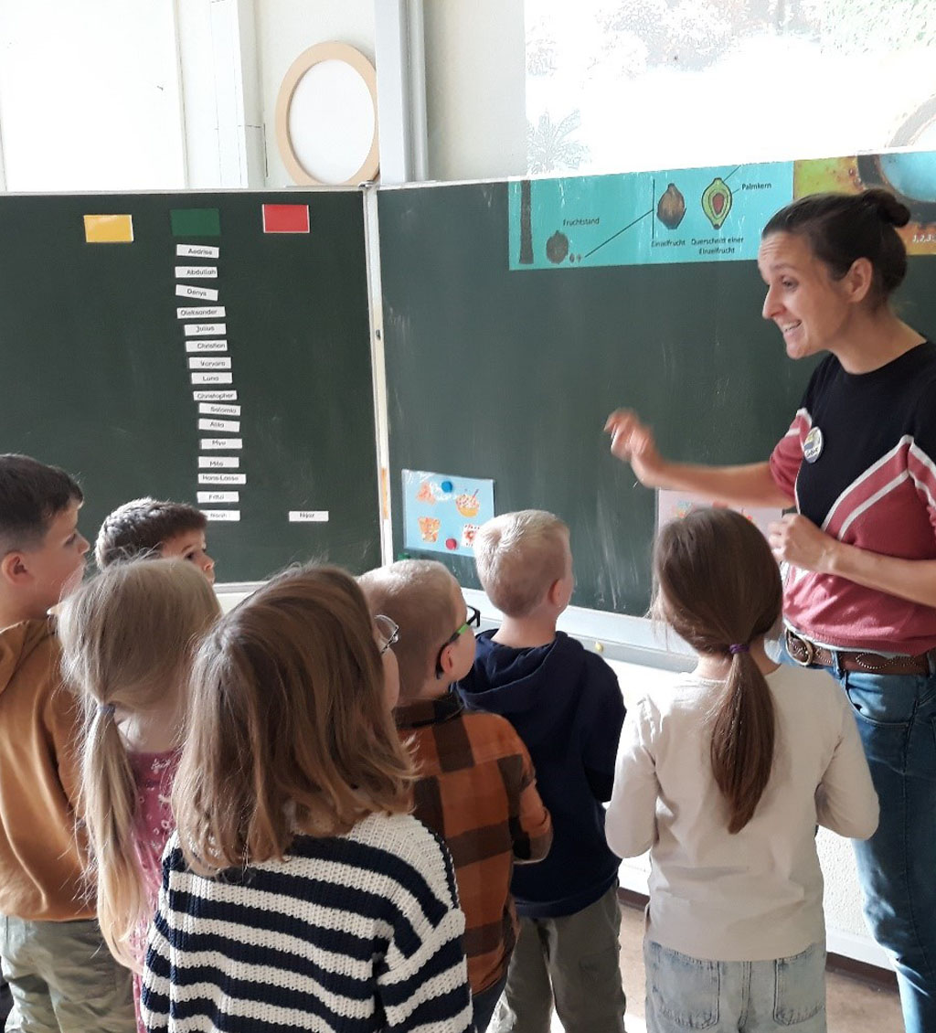 Sieben Kinder und die Umweltbildnerin stehen vor einer Tafel. An der Tafel befinden sich Bildkarten, auf denen Klebepunkte kleben.