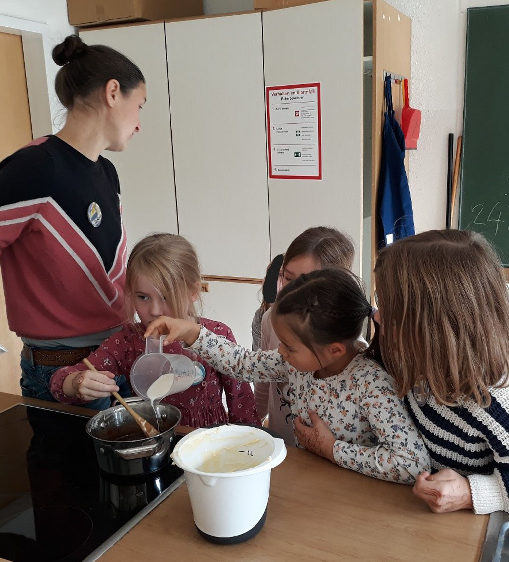 Vier Kinder und die Umweltbildnerin stehen am Herd. Ein Kind rührt in einem Topf, ein zweites gießt etwas in diesen hinein.