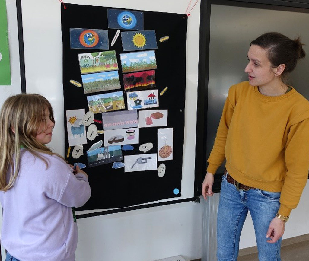 Umweltbildnerin und Schülerin stehen vor einem Tafelbild, auf dem Bildkarten zum Thema Klimawandel angeordnet sind.