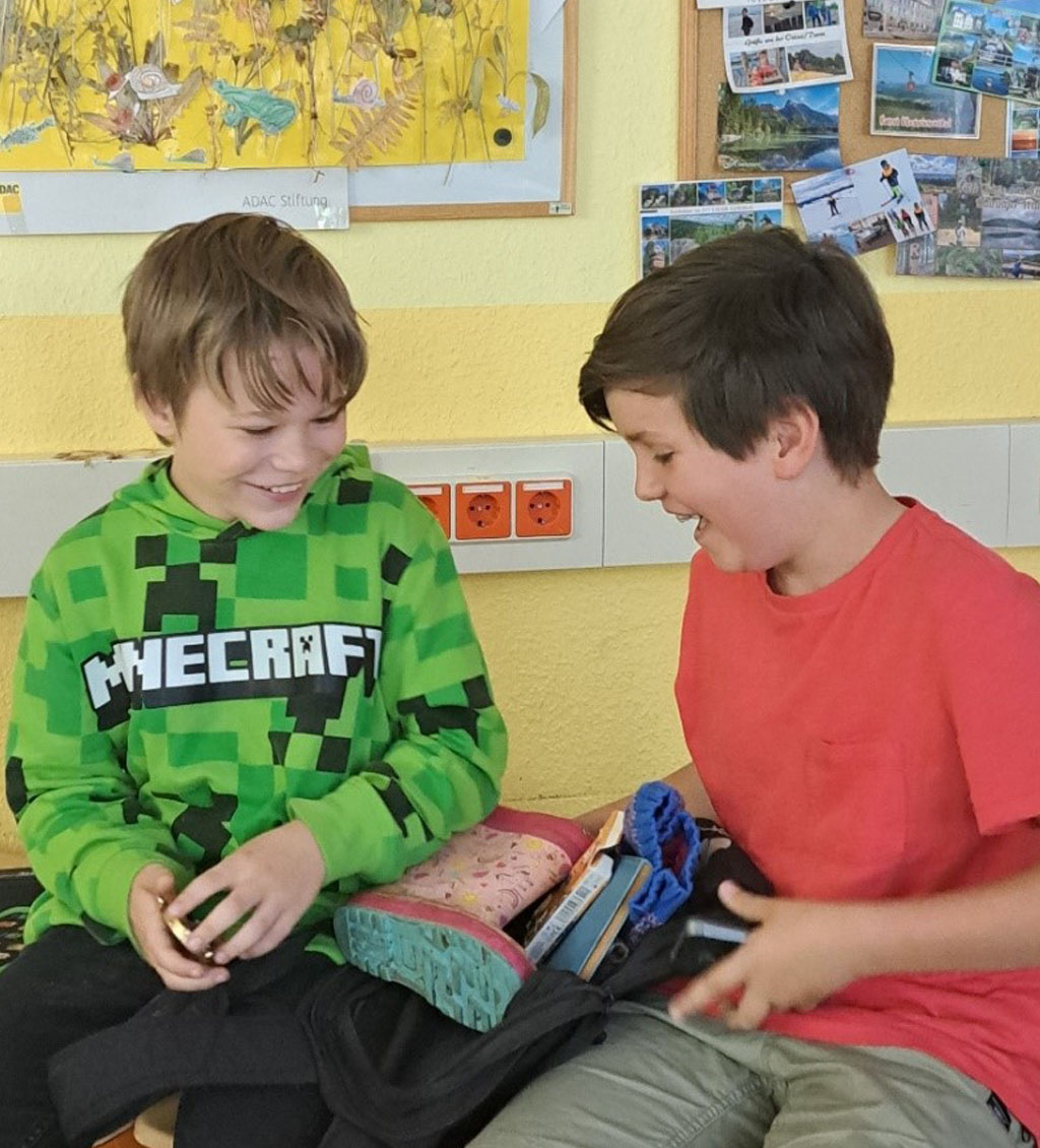 Zwei Kinder sitzen auf einem Tisch im Klassenzimmer, sie suchen Gegenstände aus einem Rucksack und lachen dabei.