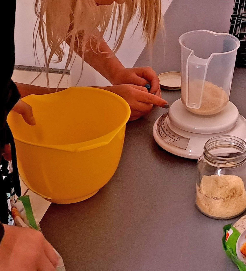 Zwei Personen stehen an einem Tisch. Darauf steht eine Waage mit Messbecher, ein Glas und eine Packung gemahlene Haselnüsse. Eine der Personen hält eine gelbe Schüssel in der Hand.