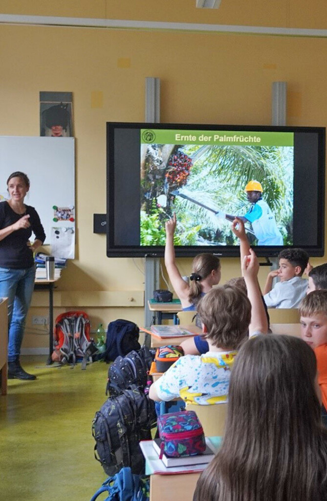 Die Umweltbildnerin steht in einem Klassenraum. An der digitalen Tafel wird eine Abbildung zur Ernte von Palmölfrüchten gezeigt. Mehrere Kinder melden sich.