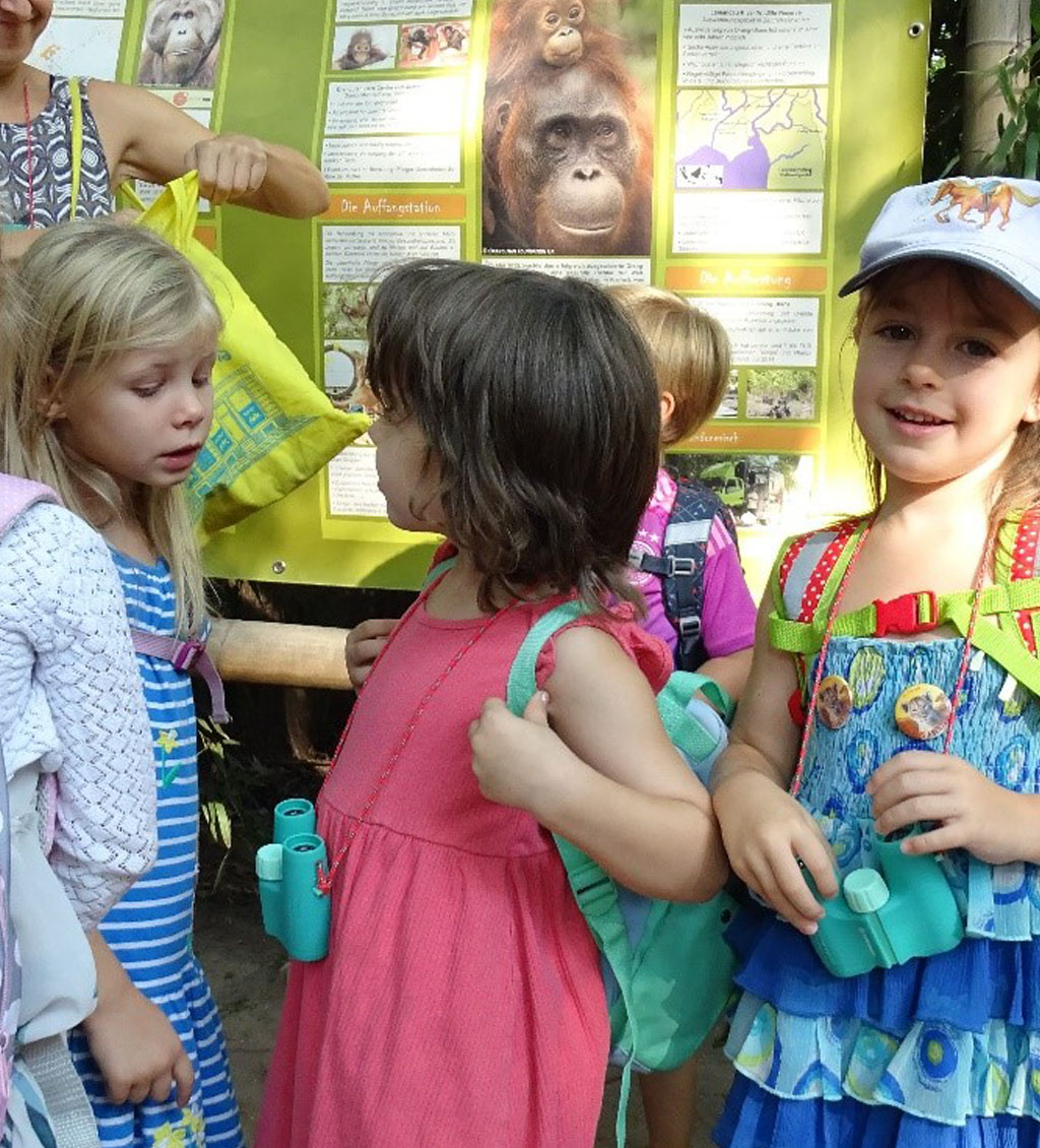 Eine Gruppe Kinder steht vor einem Infoplakat. Die Kinder tragen teilweise Ferngläser um den Hals.