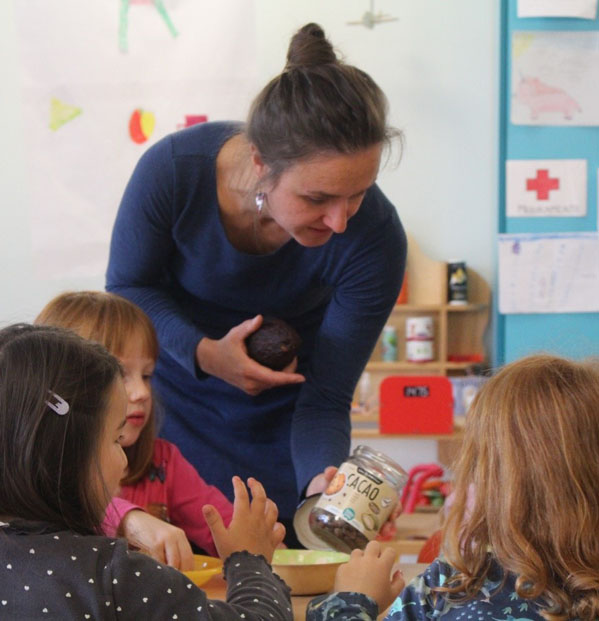 Drei Kinder sitzen an einem Tisch. Die Umweltbildnerin steht dahinter und zeigt eine Kakaofrucht und ein Glas mit Kakaobohnen.