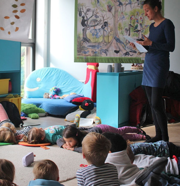 Mehrere Kinder liegen auf dem Boden, darauf befindet sich auch ein Lautsprecher. Daneben steht die Umweltbildnerin und hält ein Blatt Papier in der Hand. Hinter ihr hängt ein Regenwaldplakat.