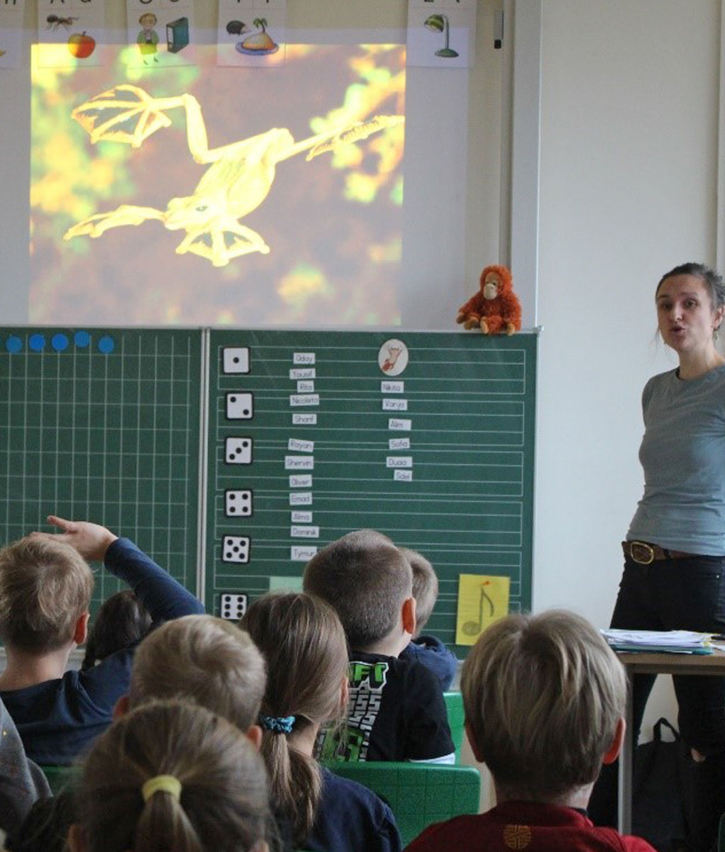 Mehrere Kinder sitzen vor einer Tafel und schauen auf eine Präsentation. Zwei Kinder melden sich. Neben der Tafel steht die Umweltbildnerin.