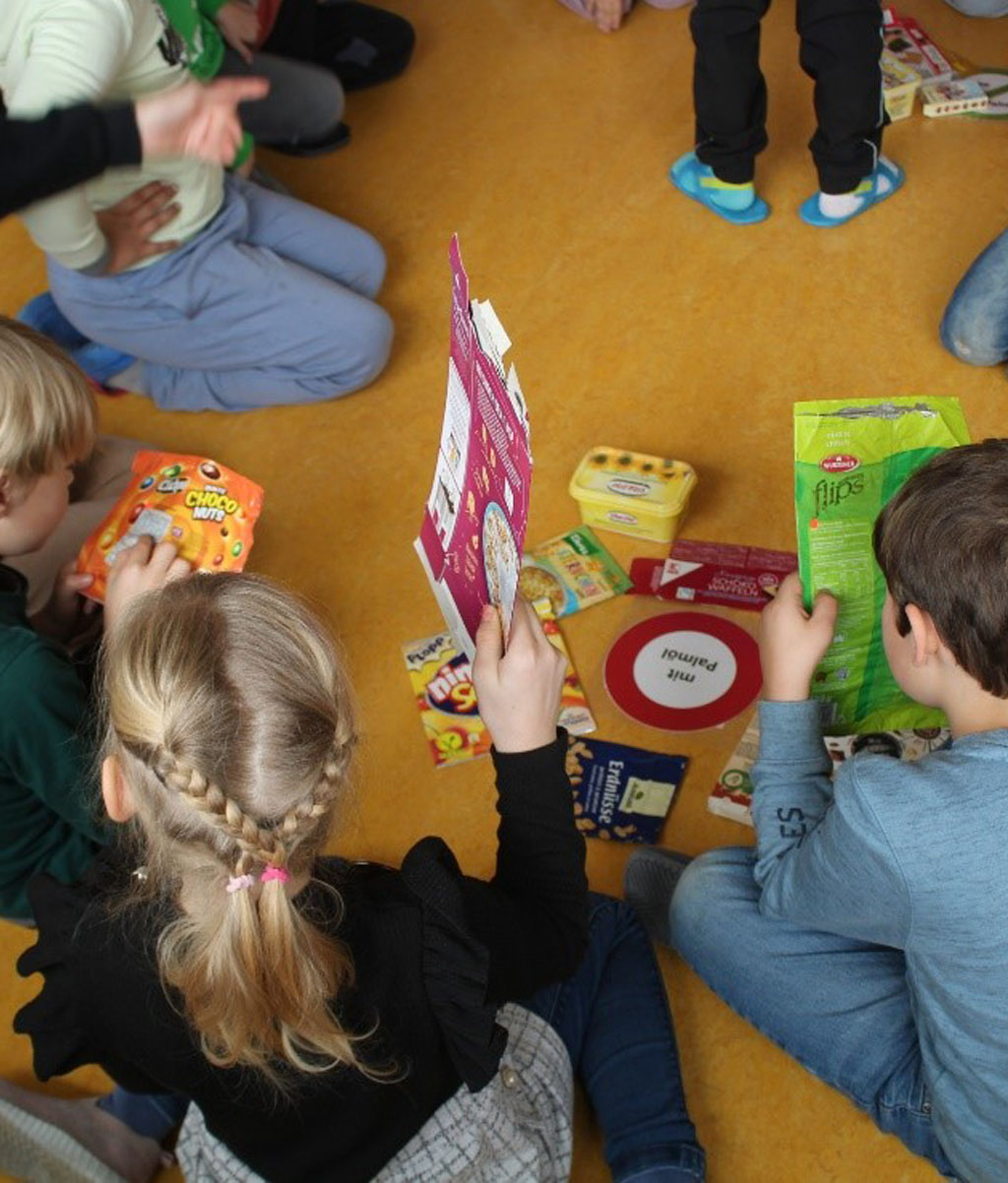 Kinder sitzen und hocken auf dem Boden. Sie halten Produktverpackungen in den Händen. Auf dem Boden liegt ein Schild mit der Aufschrift „Mit Palmöl“. Rings um das Schild liegen weitere Verpackungen.