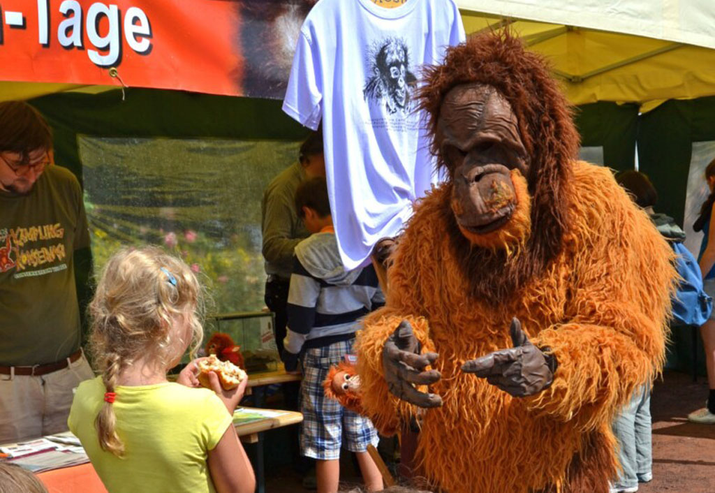 Der Kostüm-Orang-Utan redet vor einem Informationsstand mit einem Kind.